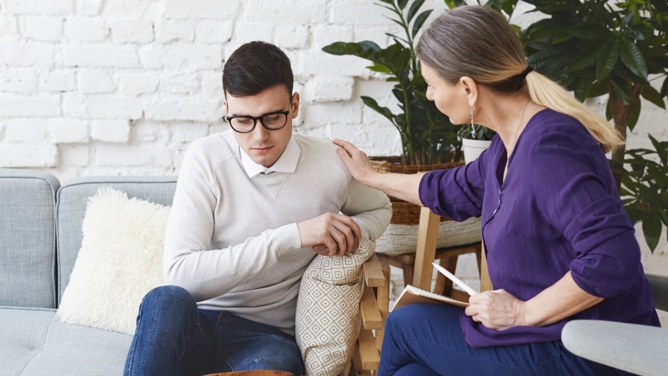 Candid shot of casually dressed professional woman psychotherapist in her fifties touching her young male patient by shoulder while having counseling session, expressing sympathy and support